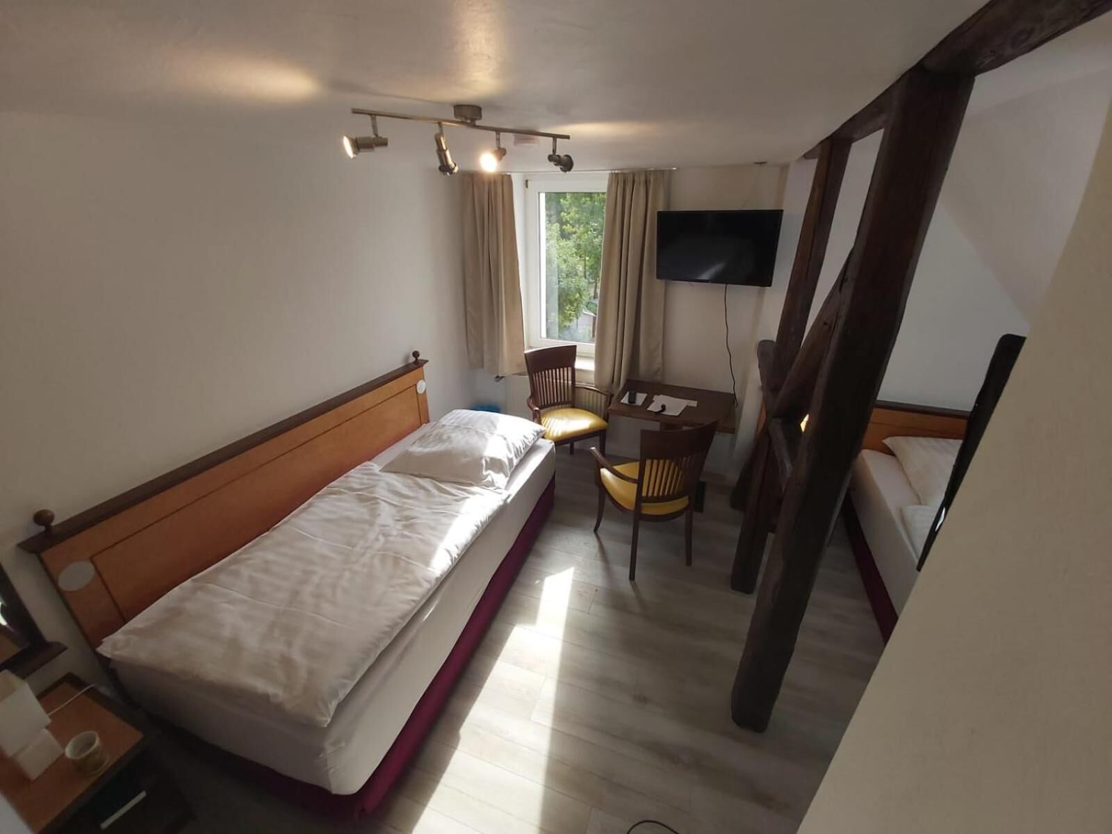 A hotel room with a bed, a desk with a chair, a TV mounted on the wall, and a window with curtains.
