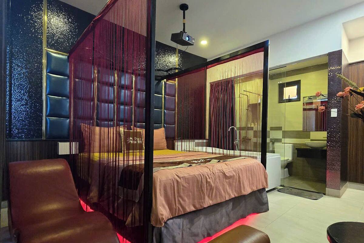 Un dormitorio moderno con cama con dosel, un rincón de estar con una silla y un baño visible a través de un mamparo de vidrio.