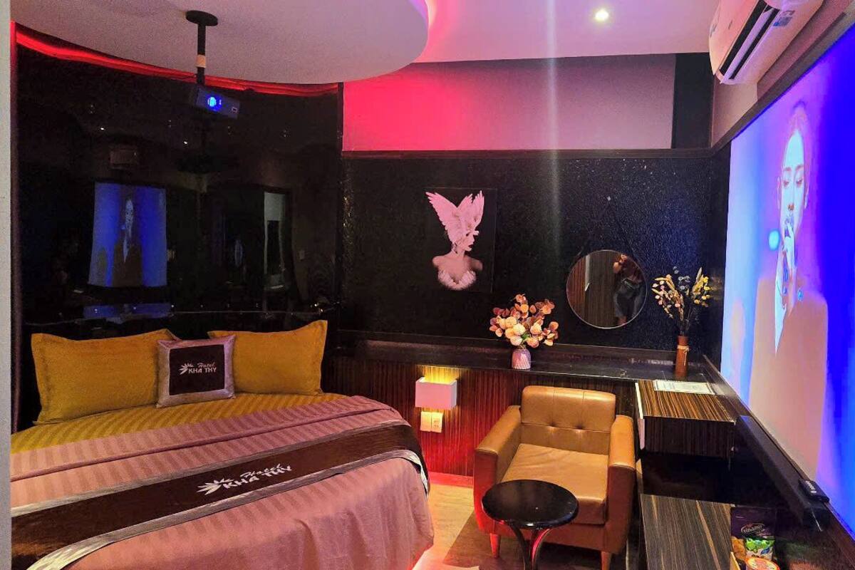 Habitación de hotel con cama, una silla, una mesita, un espejo y una pantalla de televisión que muestra a una persona.