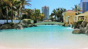 A clear blue pool with a bridge, surrounded by palm trees and rocks, with a beach in the foreground and a tall building in the background.