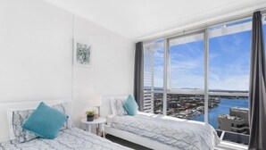 A hotel room with two beds, a small table with a lamp, and a large window offering a view of a city and water.