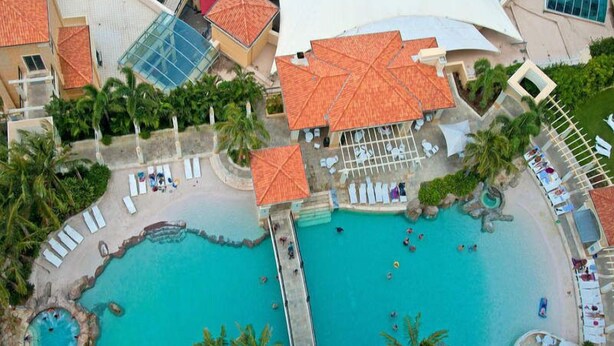 An aerial view of a resort with two large swimming pools, a beach area, and a covered pavilion.