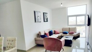 Un salon moderne avec un canapé beige, un fauteuil rose et une table basse en verre. Deux œuvres d’art encadrées ornent le mur, et un téléviseur y est fixé.