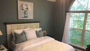 A bedroom with a bed, pillows, a nightstand, a lamp, and a window with a view of trees.