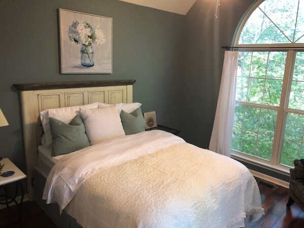 Une chambre à coucher avec un lit, des oreillers, une table de chevet, une lampe et une fenêtre donnant sur des arbres.