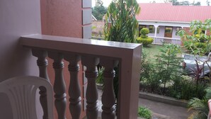 Un balcon doté d’une balustrade blanche, donnant sur un jardin où poussent différentes espèces de plantes, avec, au loin, un bâtiment au toit rouge.