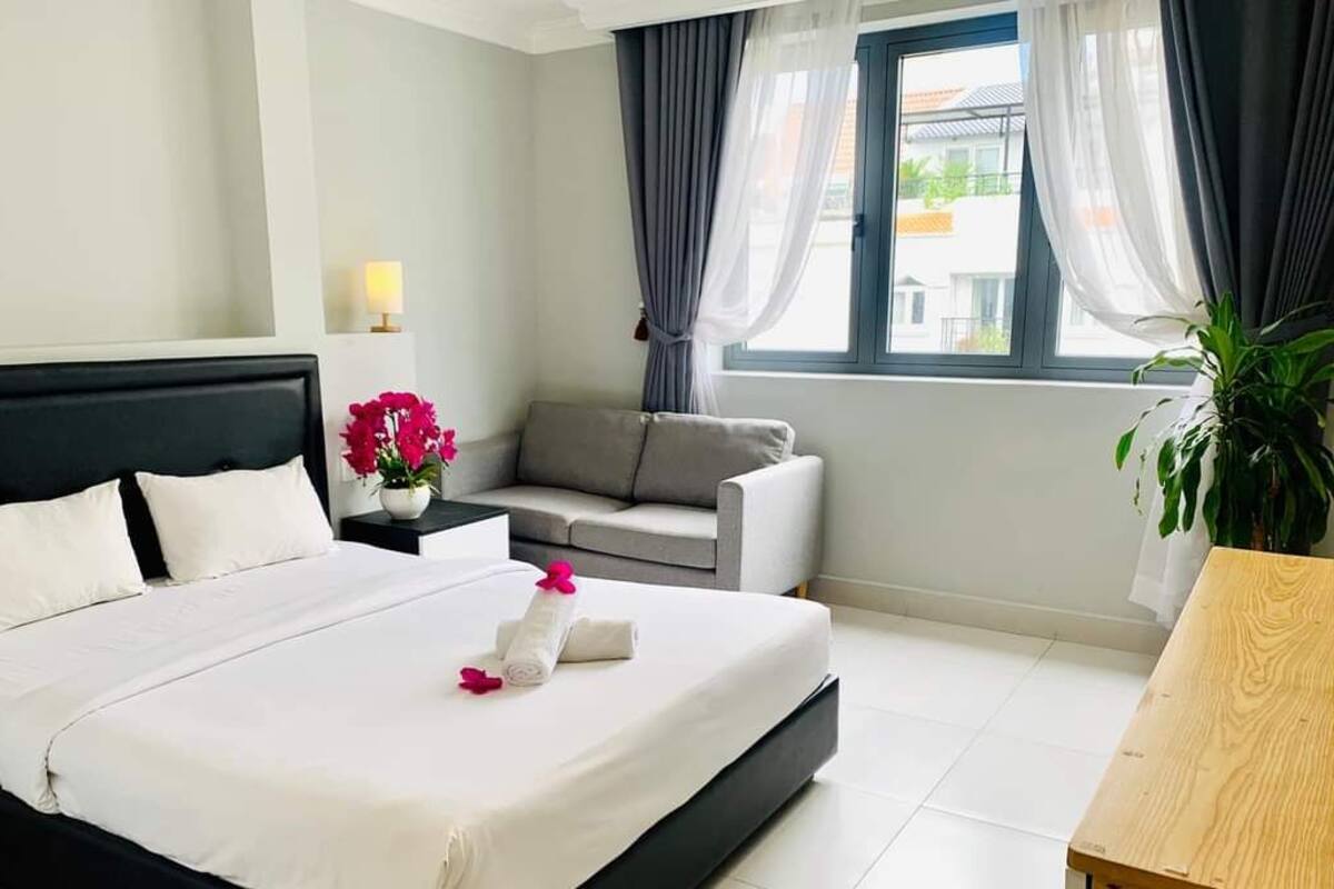 A hotel room with a bed, a grey sofa, a small table with a plant, and a window with curtains.