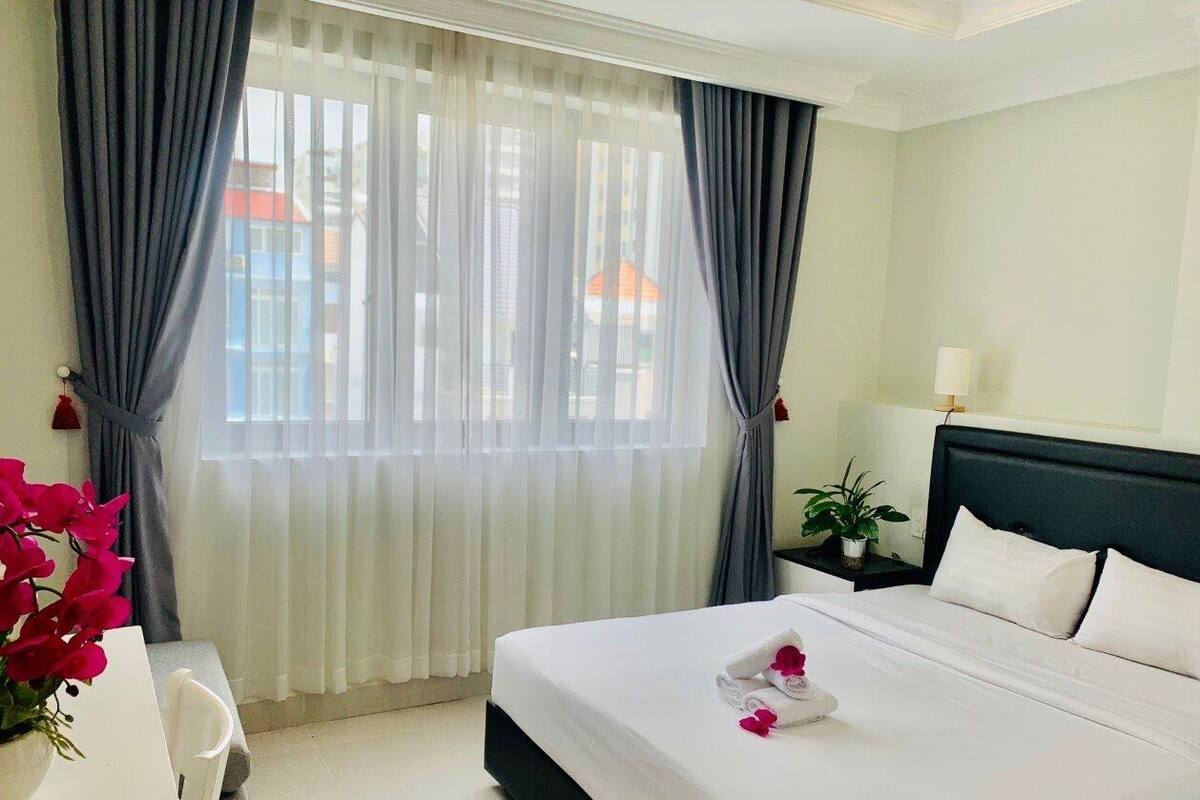 A hotel room with a bed, a nightstand, a lamp, a window with curtains, and a view of buildings outside.