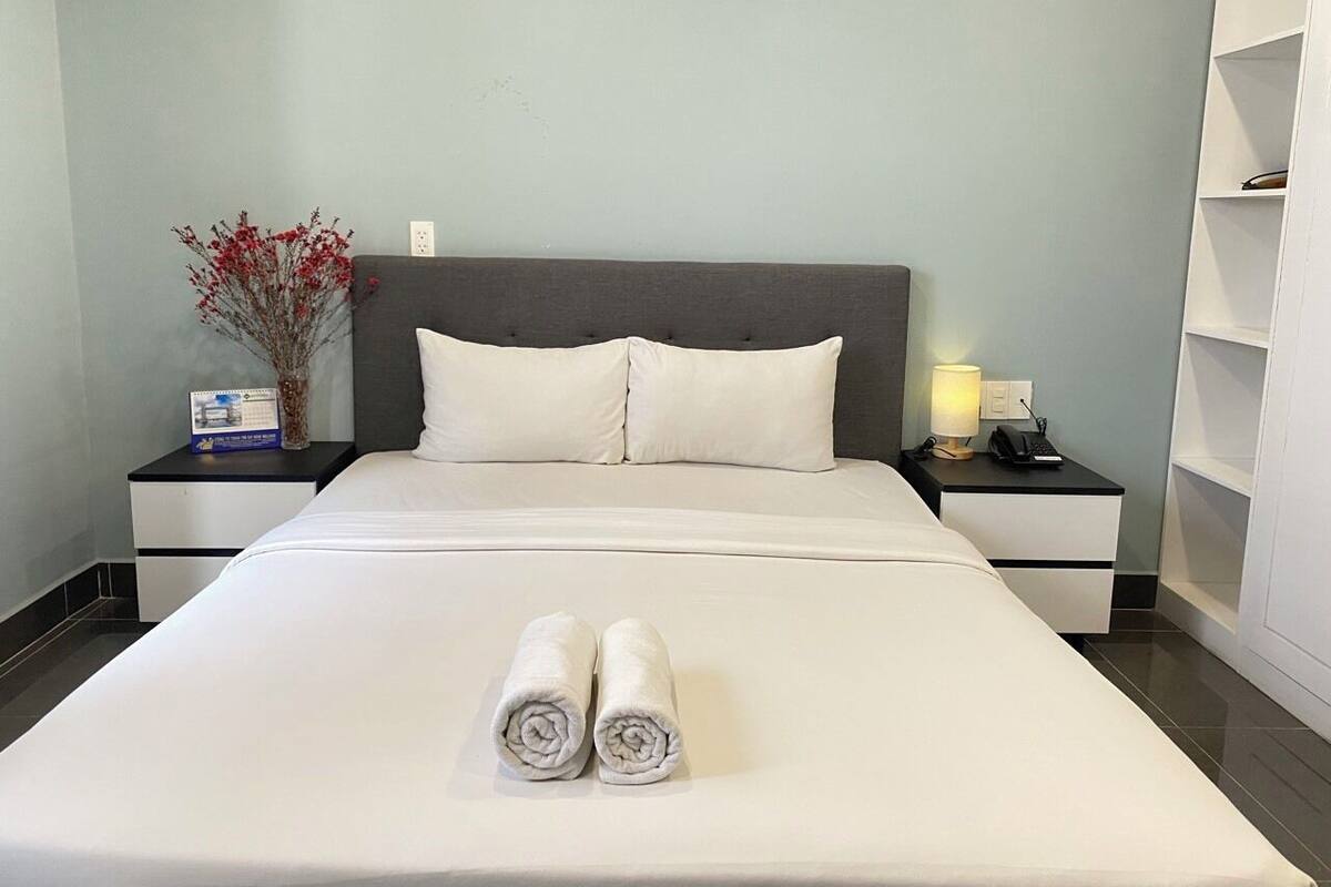 A neatly made bed with two rolled towels, a nightstand with a lamp and phone, and a vase with red flowers on the left side.
