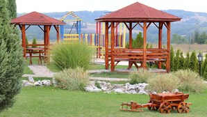 Two wooden gazebos with benches, surrounded by grass and rocks, with a playground and mountains in the background.