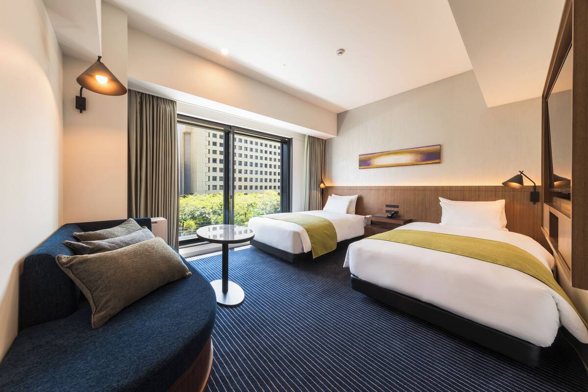 Een hotelkamer met twee bedden, een bank, een tafeltje en een groot raam met uitzicht over de stad.