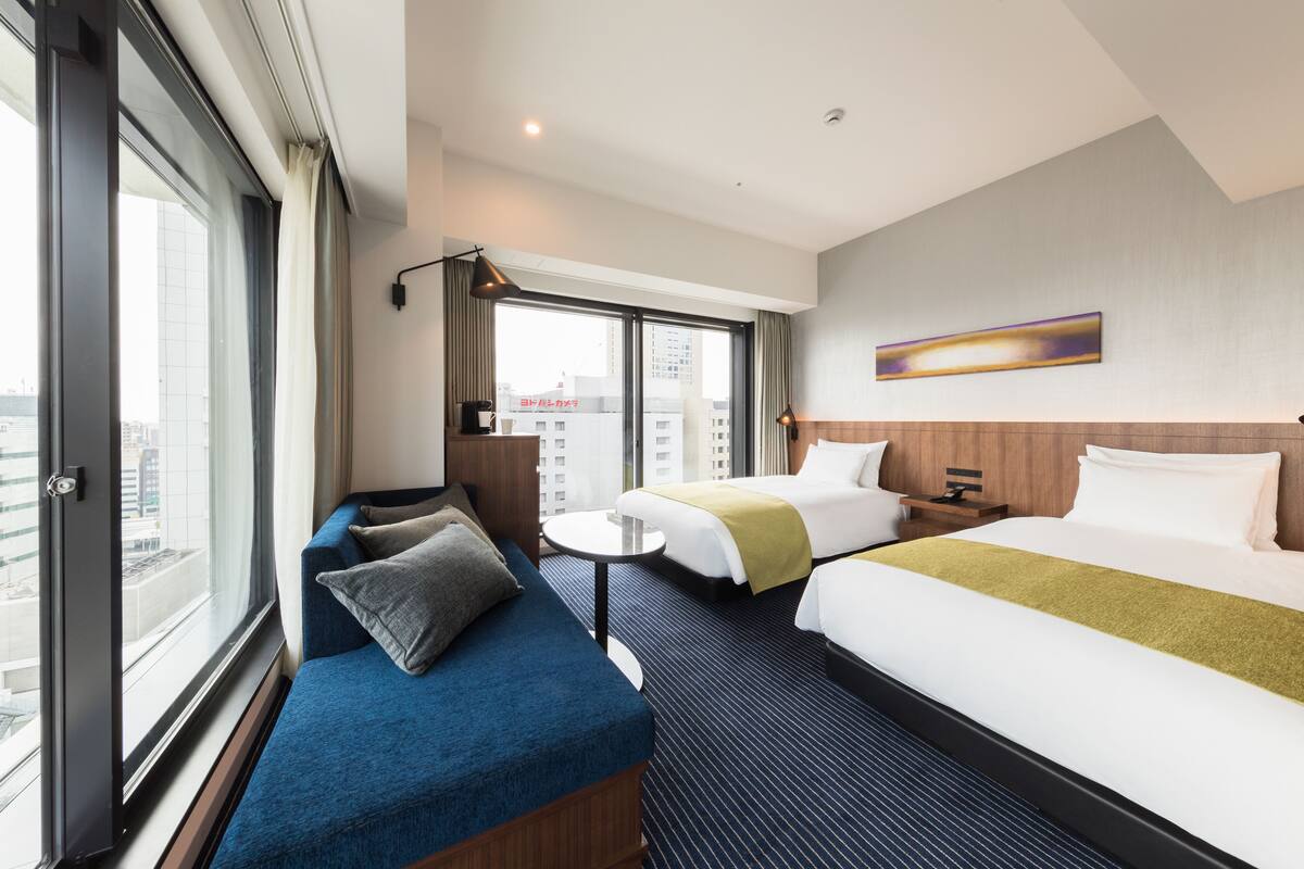 Een hotelkamer met twee bedden, een bank, een kleine tafel en een groot raam met uitzicht op de stad.