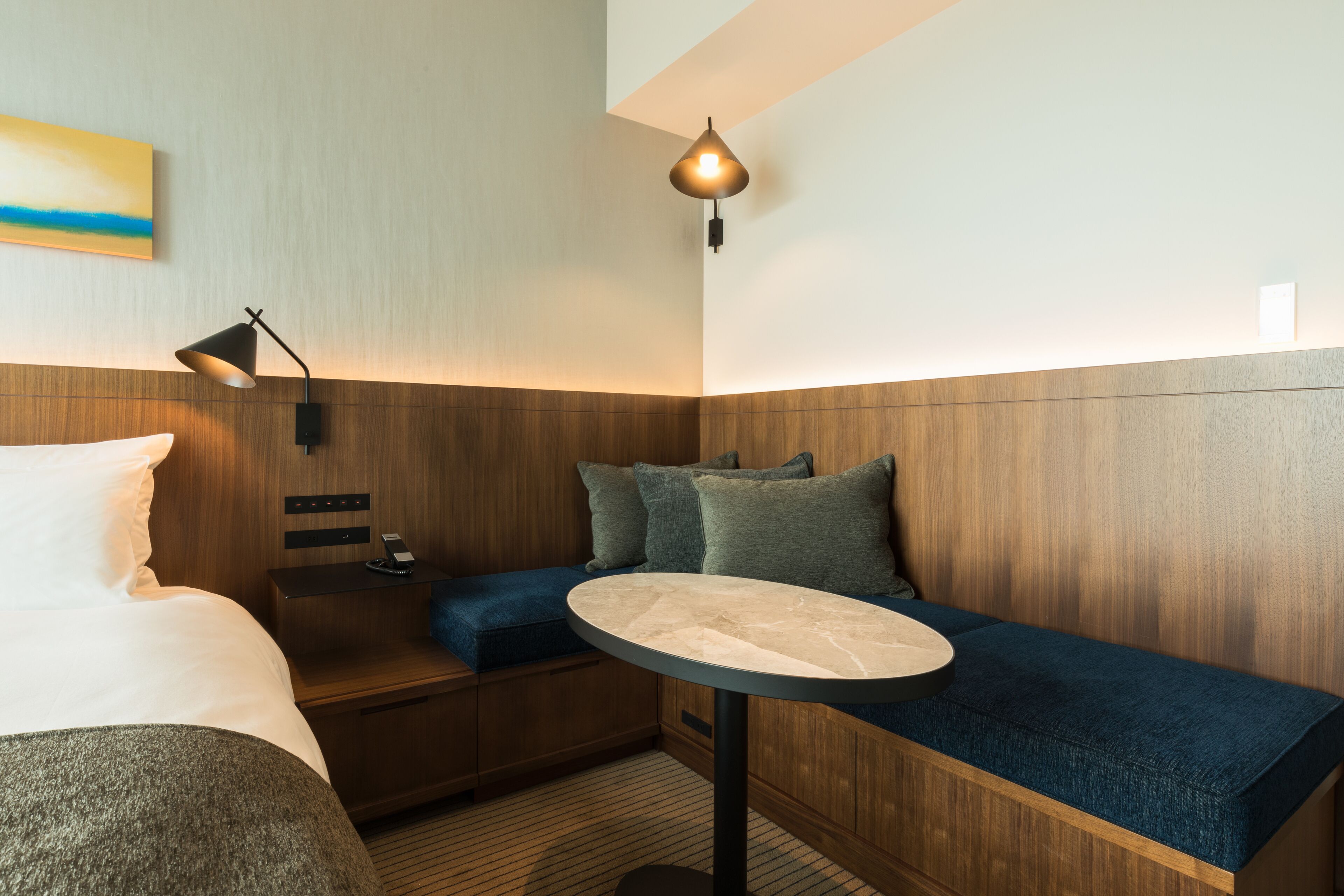 Habitación de hotel moderna con cama, una mesita redonda, un banco y una lámpara de pared.