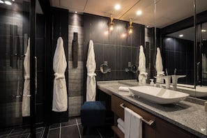Une salle de bain moderne avec des carreaux foncés, un grand miroir, un lavabo blanc et deux serviettes blanches suspendues à des crochets.