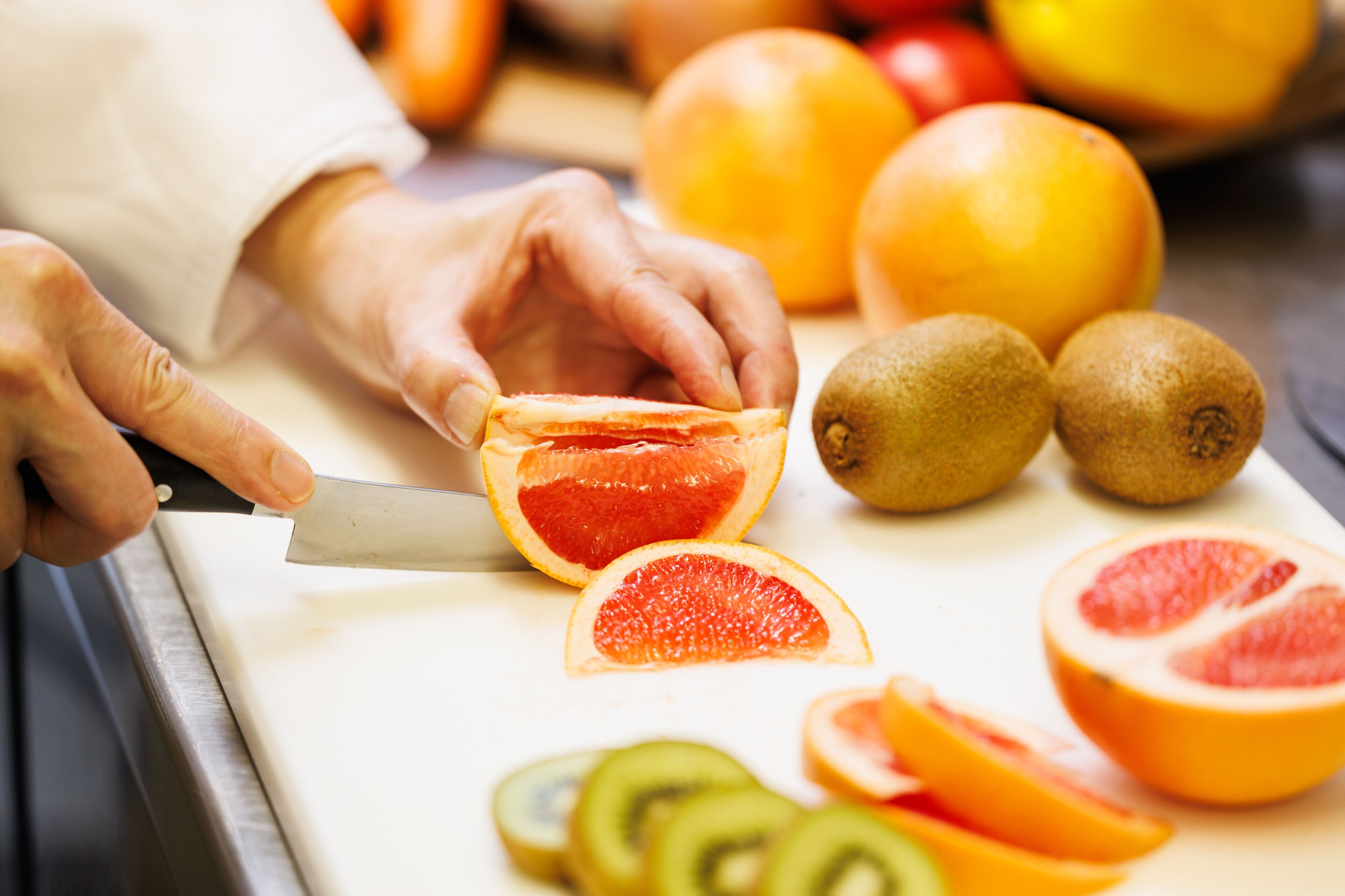 A person slicing a grapefruit on a cutting board with other fruits like kiwi and oranges nearby.