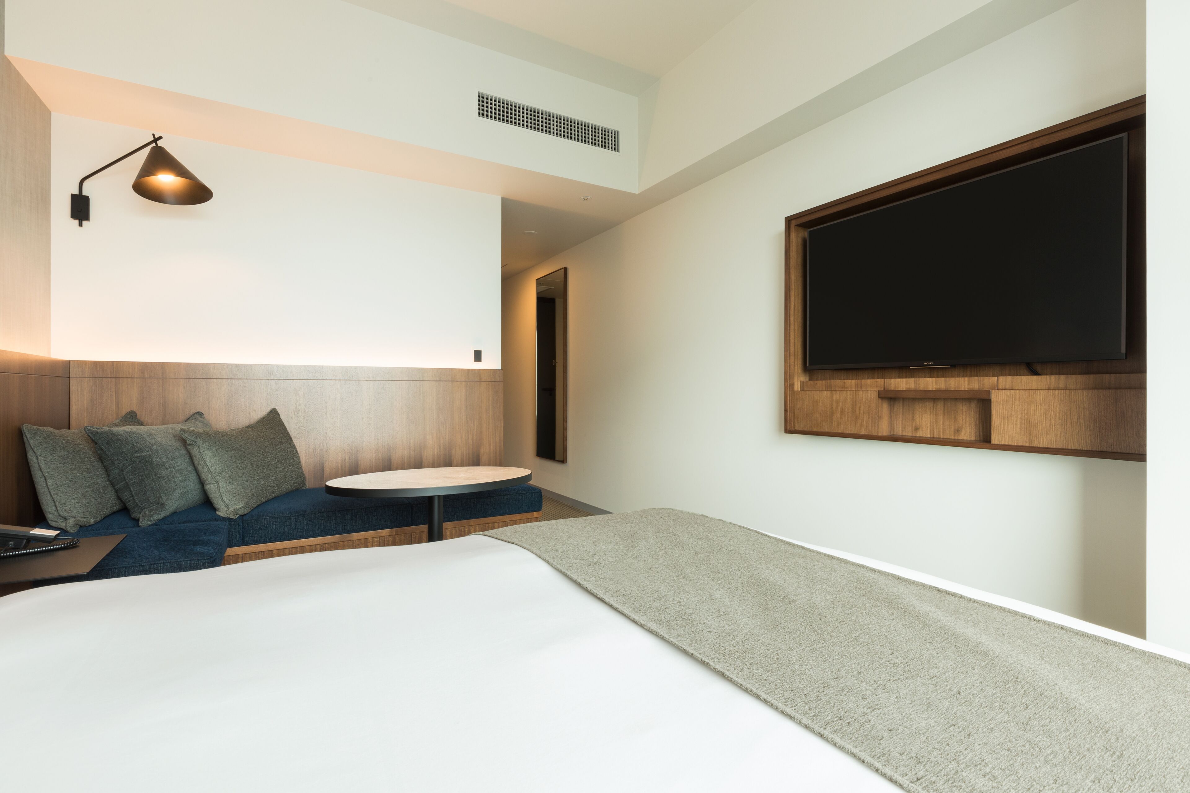 A modern hotel room with a bed, a desk, a chair, a small table, and a TV mounted on the wall.