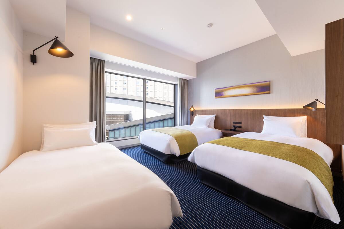 Een hotelkamer met twee bedden, een groot raam met uitzicht op gebouwen, een moderne lamp en een abstract kunstwerk aan de muur.