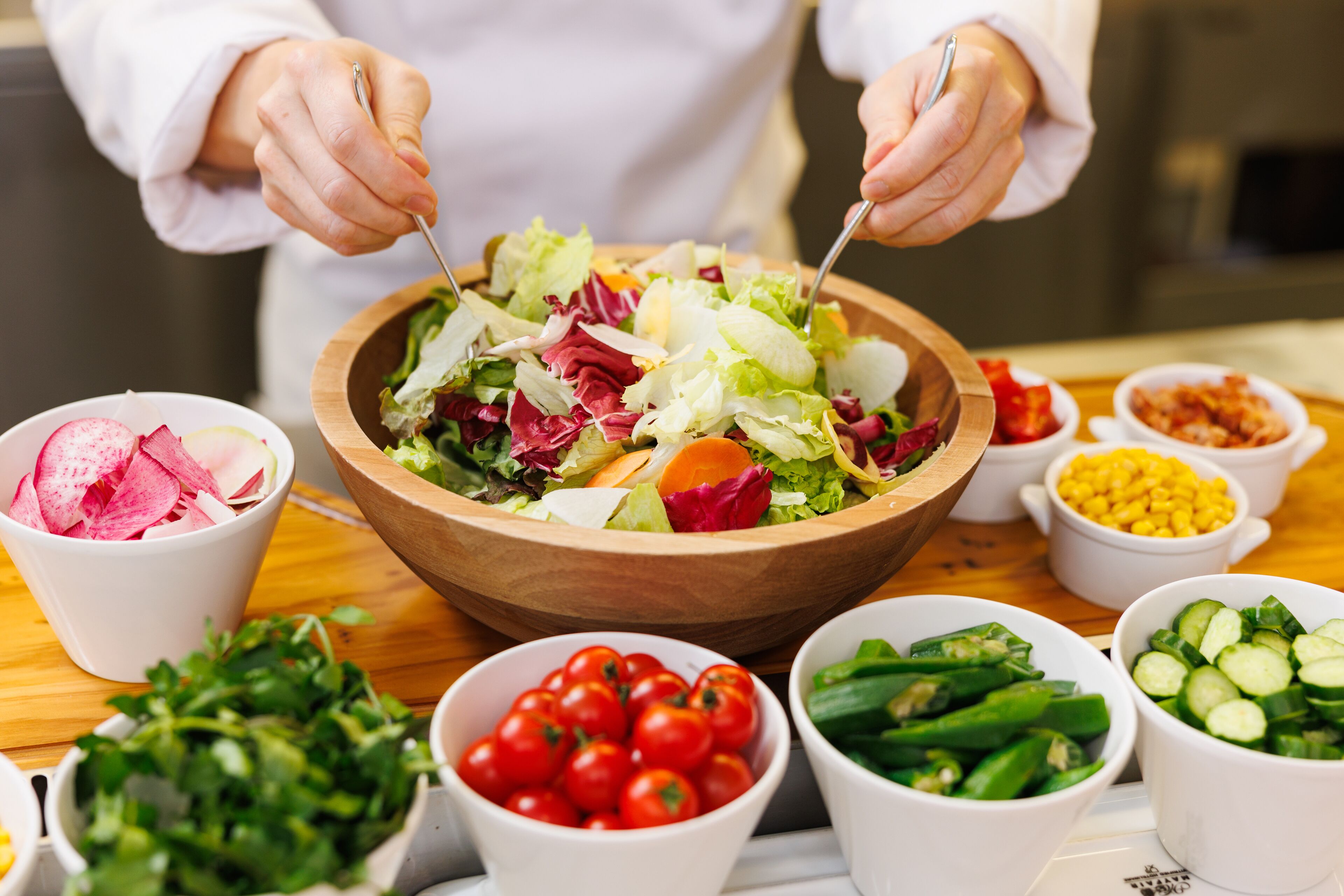 Un chef preparando una ensalada con diversas verduras y hojas verdes en un cuenco de madera.