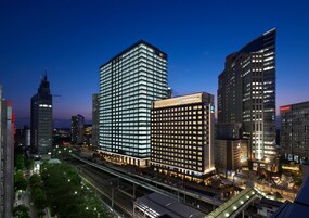 Un hotel moderno con una fachada de vidrio destacada, rodeado de edificios altos y una concurrida calle urbana al anochecer.
