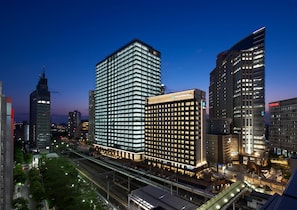 Un hôtel moderne à la façade imposante, entièrement vitrée, entouré de gratte-ciel et bordé d’une rue animée, le tout baigné par la lumière du crépuscule.