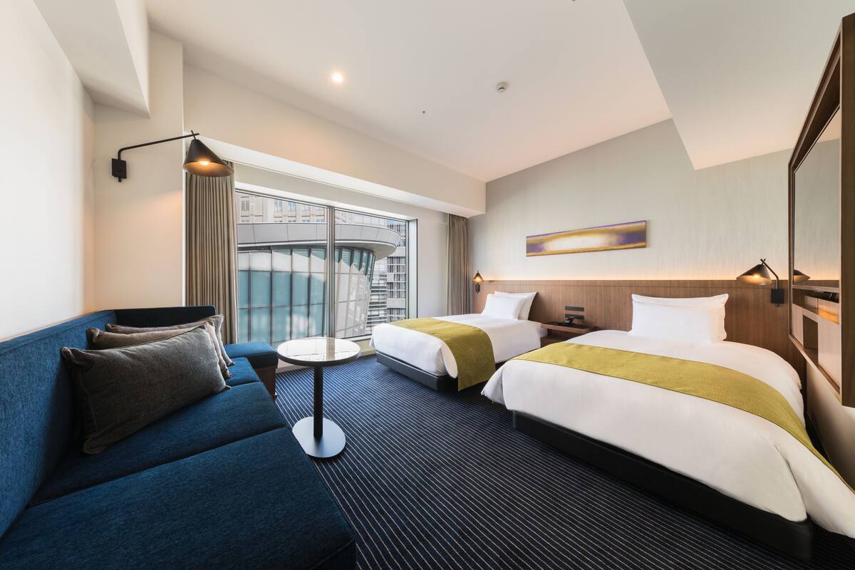 Een hotelkamer met twee bedden, een sofa, een kleine tafel en een groot raam met uitzicht op een gebouw.