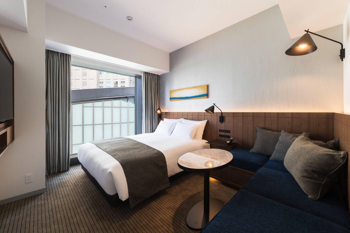 Een hotelkamer met een bed, een bank, een kleine tafel, een raam met gordijnen en een schilderij aan de muur.