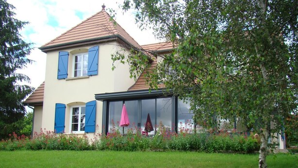 A two-story house with a sloped roof, large windows, and a small garden area with flowers and a tree. - Chambres sous les vignes du Buttois (Bussy-le-Grand)