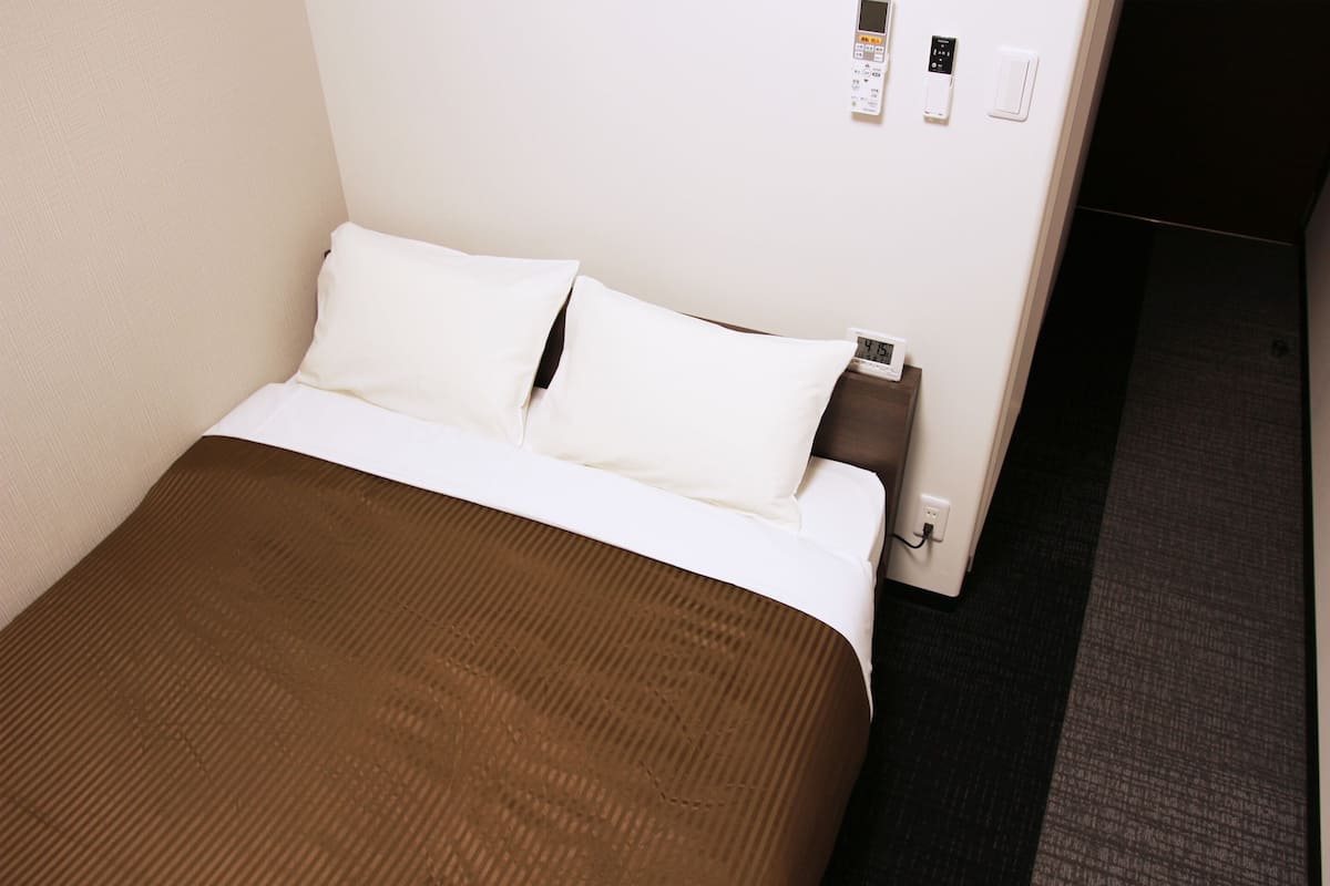 Cama bem arrumada com lençóis brancos e manta marrom, mesa de cabeceira com telefone e tomada, e termostato fixado na parede.