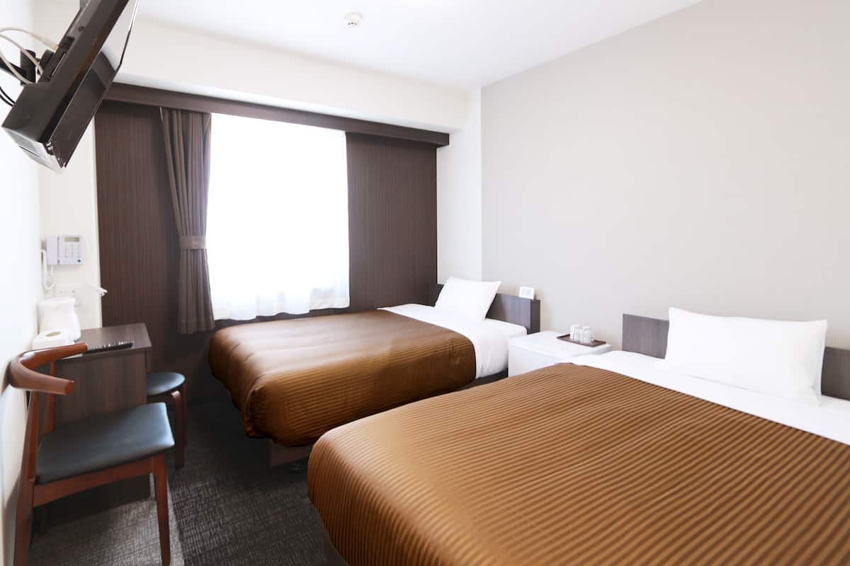 Quarto de hotel com duas camas, uma escrivaninha, uma cadeira, televisão e janela com cortinas.