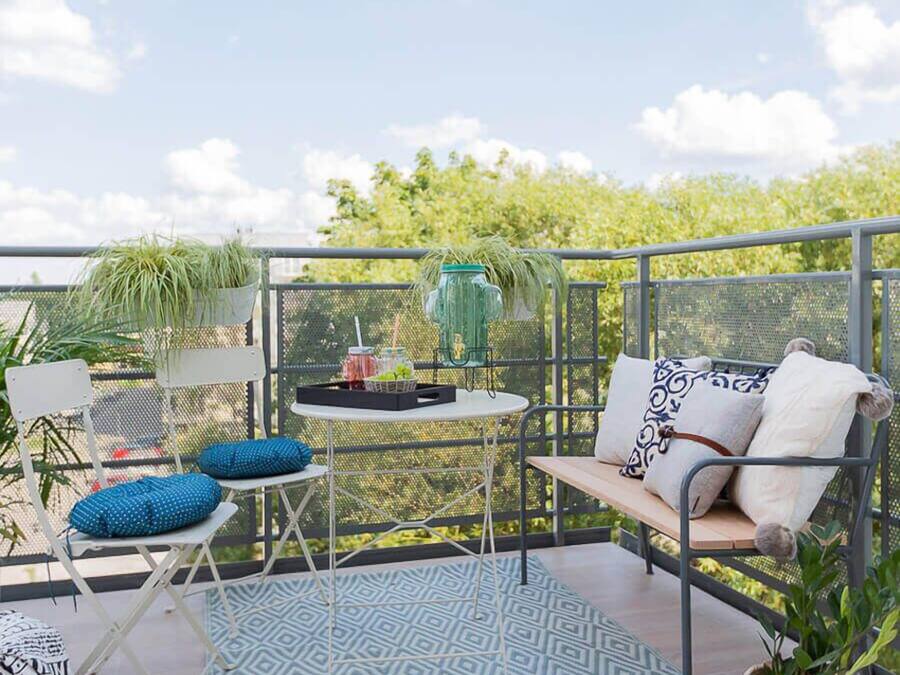 A balcony with a metal railing, a small table with drinks, a bench with cushions, and a patterned rug.