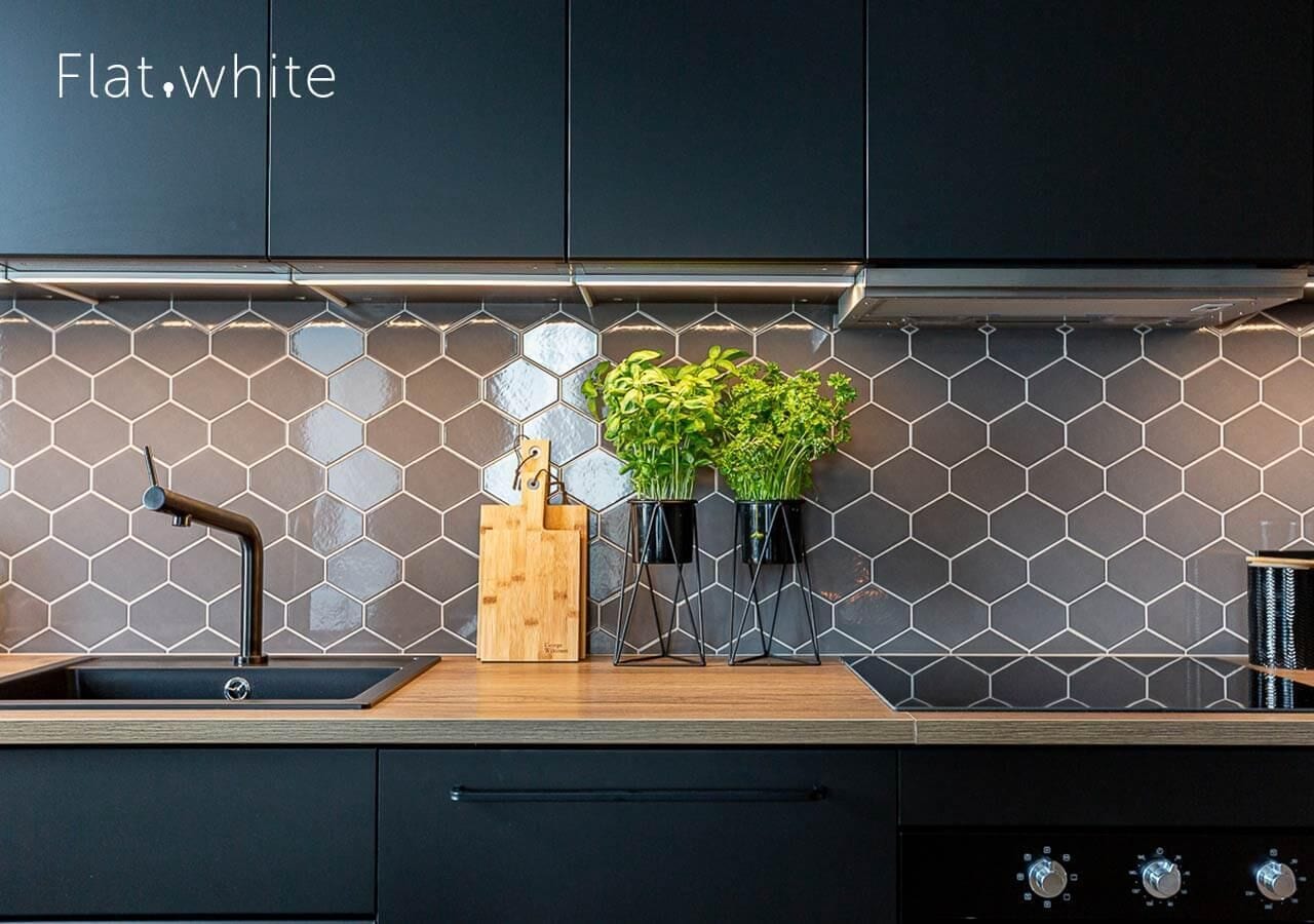A modern kitchen with a black countertop, a sink with a black faucet, and a hexagonal tile backsplash. There are two potted plants on the countertop.