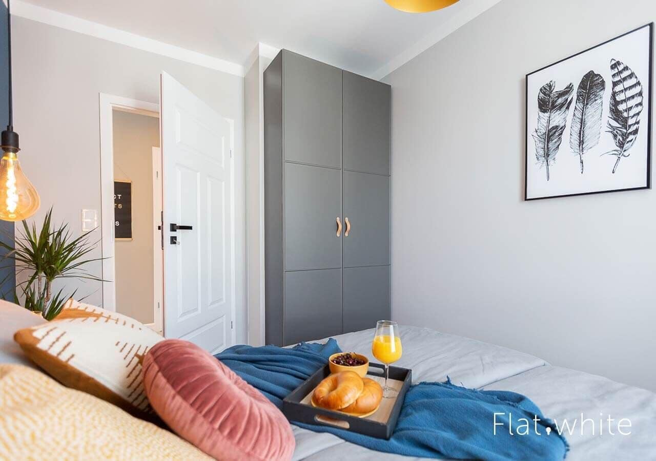 A bedroom with a bed, pillows, a tray with croissants and orange juice, a grey wardrobe, and a framed feather artwork on the wall.