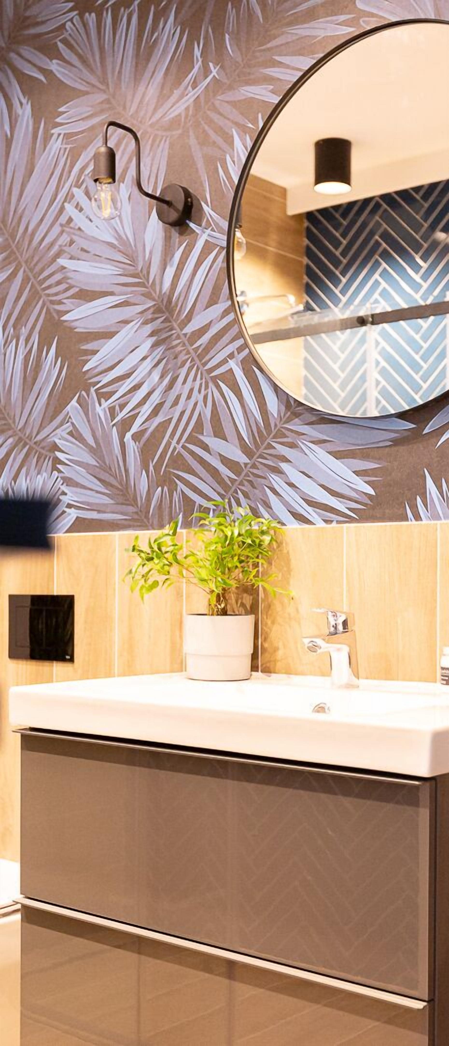 A modern bathroom with a wooden vanity, a potted plant, a round mirror, and a wall with a leaf pattern.