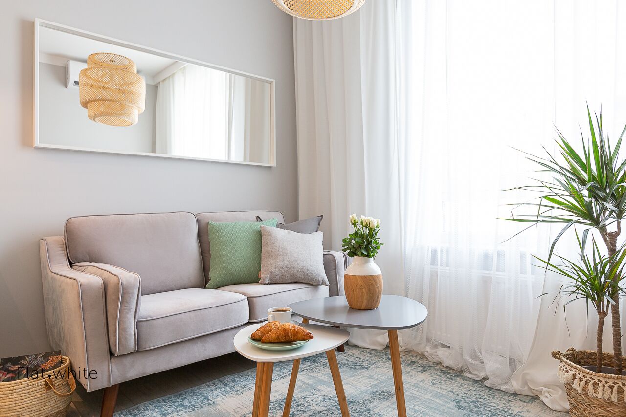 A modern living room with a grey sofa, a small round table, a potted plant, and a large mirror on the wall.
