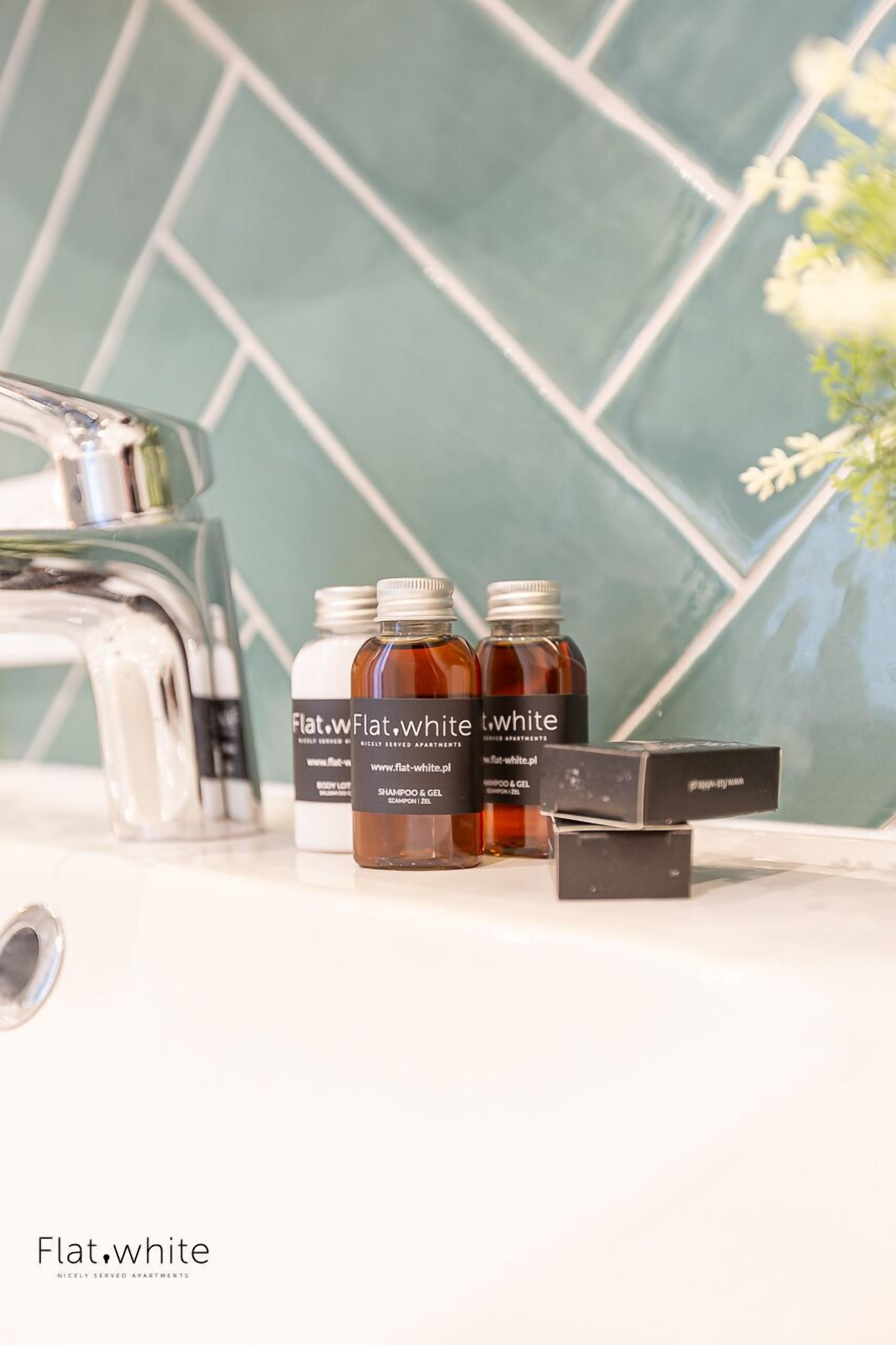 Et badeværelse med et moderne design, med et brusebad i glas, en hvid vask og en flisebeklædt væg. Der er tre flasker Flat White shampoo og gel, samt en sort æske på bordet.