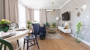 A modern living room with a dining area, a sofa, a TV mounted on the wall, and a large window with curtains. - Flat White Apartamenty Obywatelska 47 (Zielona Gora)