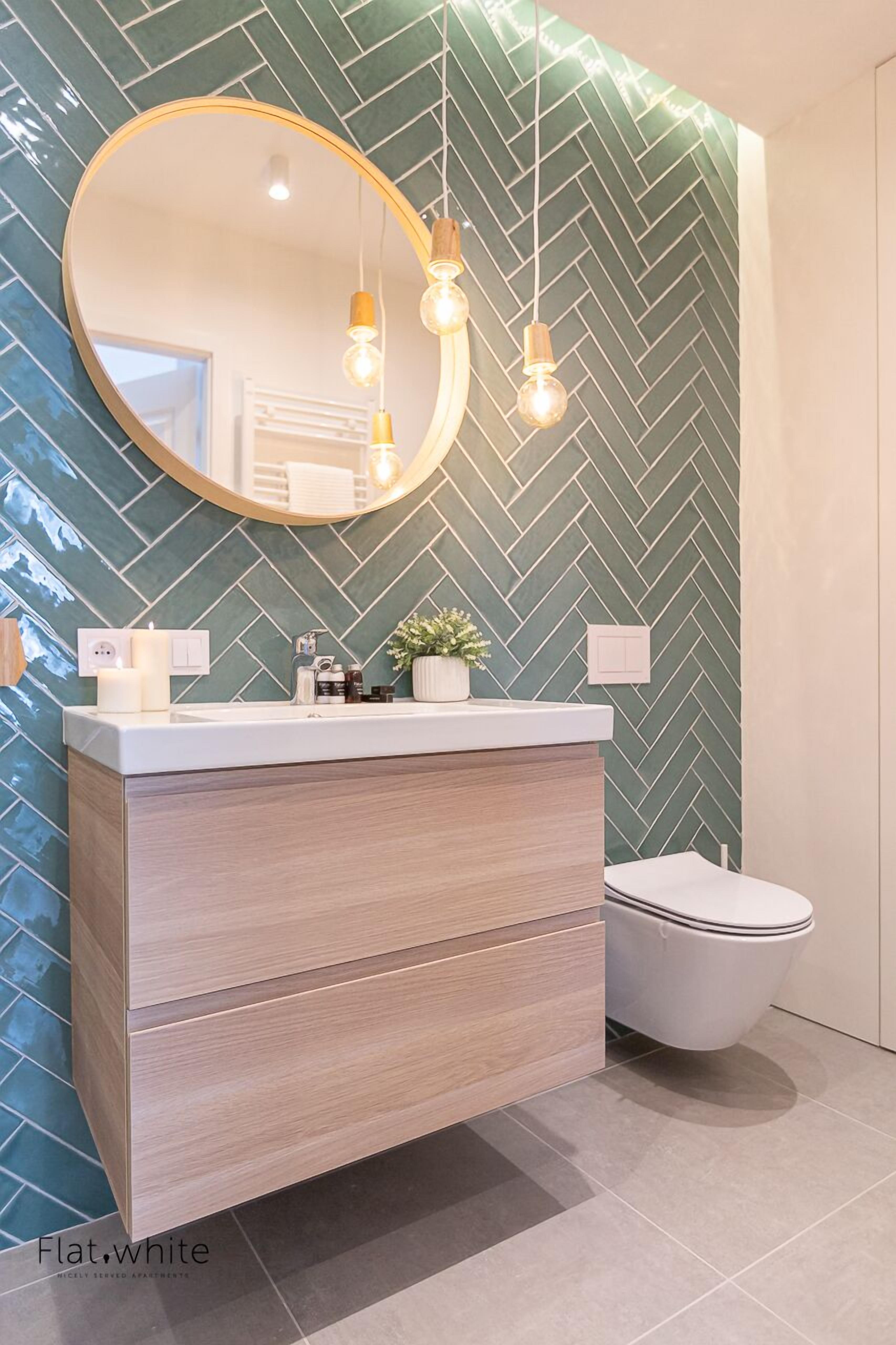 Une salle de bain avec un mur orné d’un motif en chevrons, un miroir rond, un meuble-lavabo en bois et des toilettes blanches.