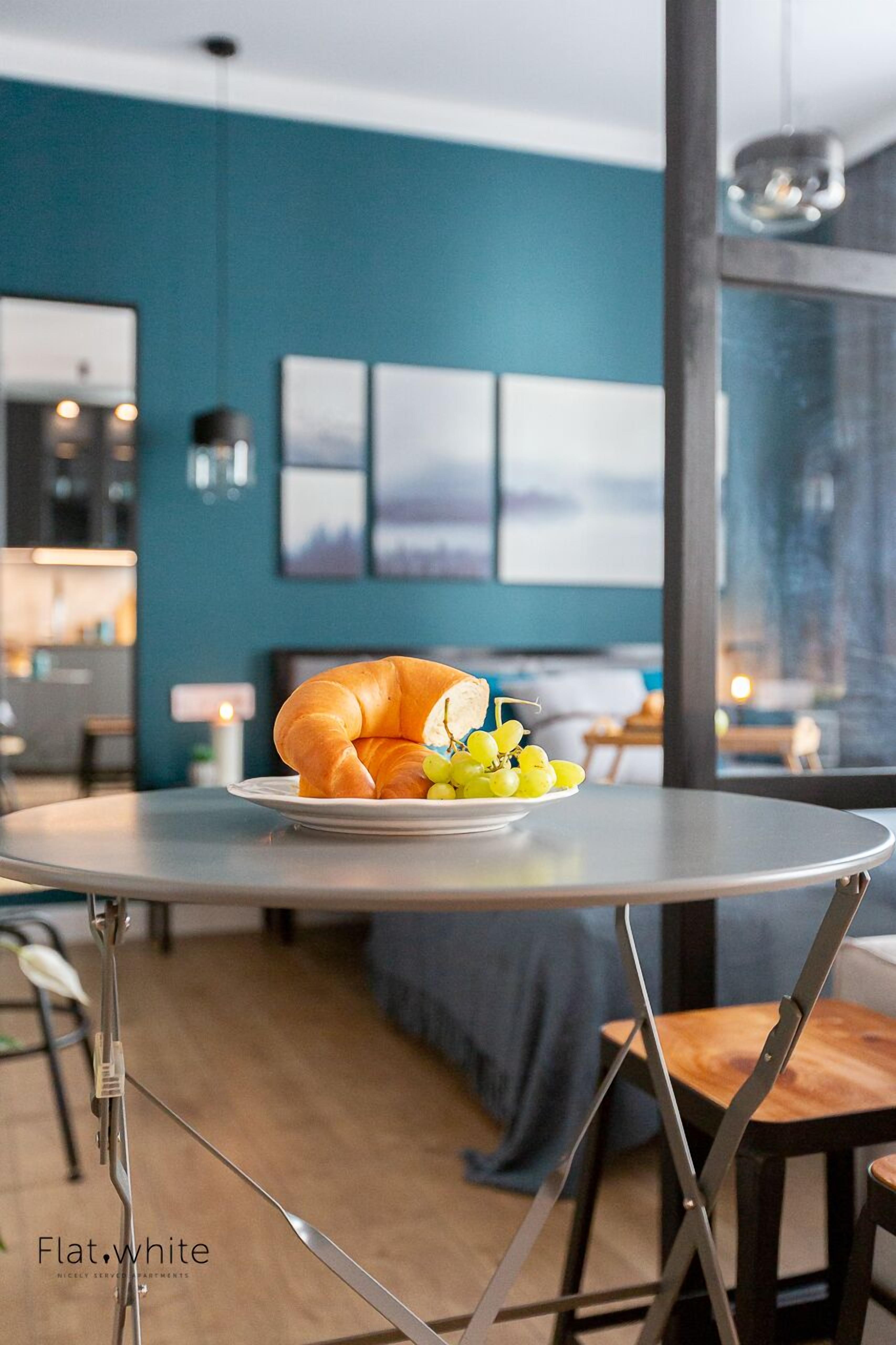 A dining table with a croissant and grapes, a modern kitchen in the background, and a view of the city through large windows.