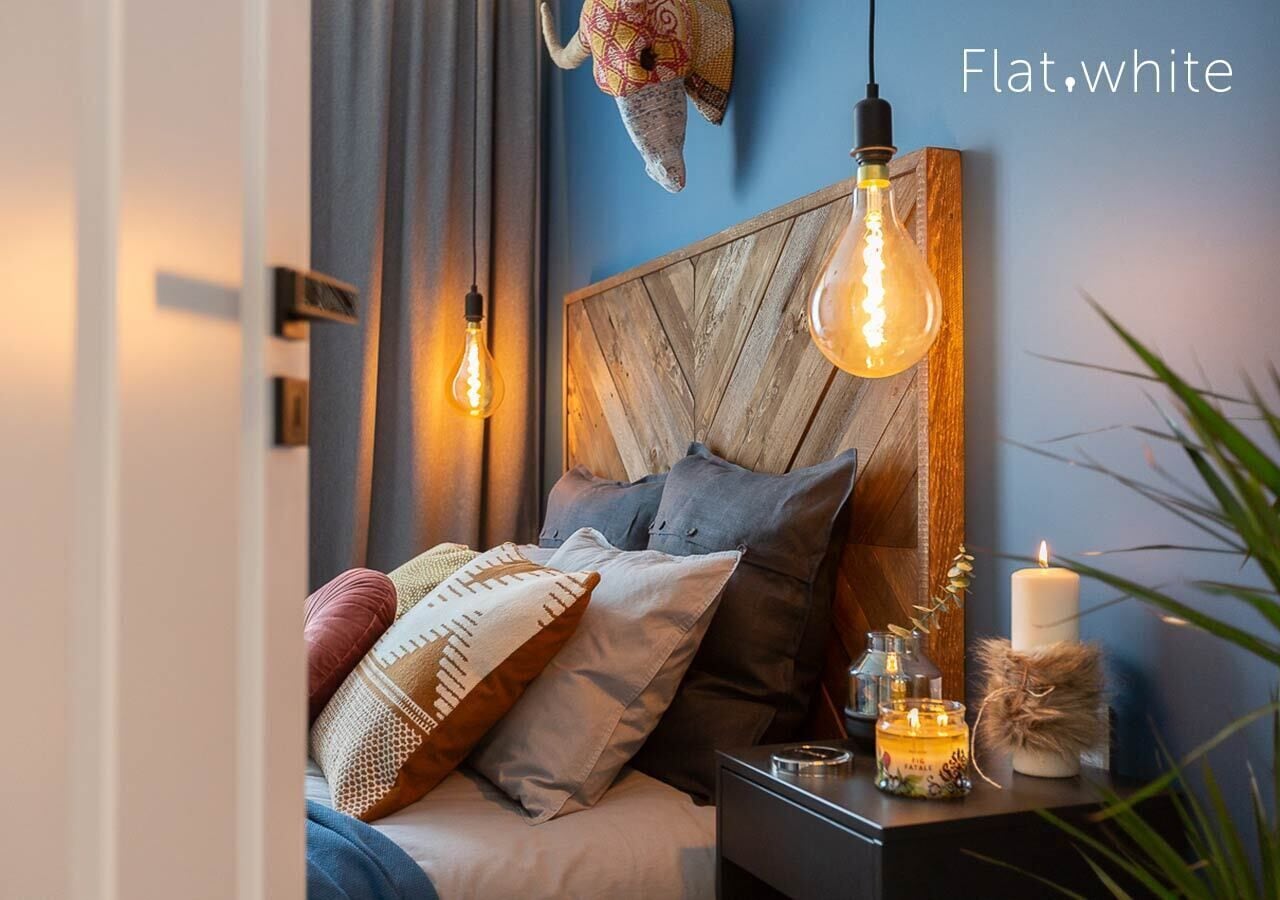 A bedroom with a wooden headboard, a bedside table with a candle, and a wall-mounted light fixture.