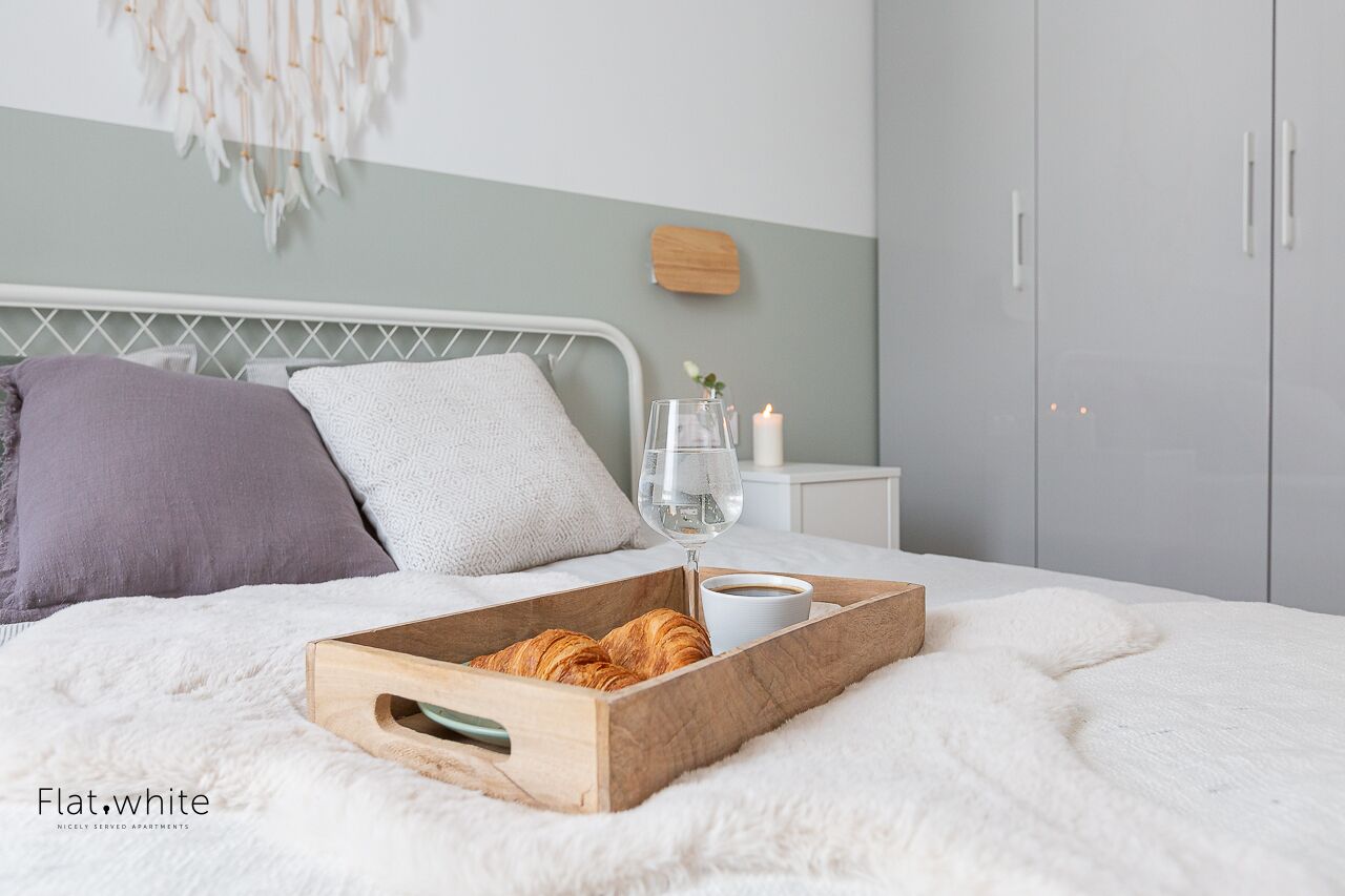 Cama bem arrumada com uma bandeja de croissants, um copo de água e uma xícara de café sobre um cobertor branco e fofo.