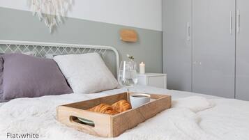 Cama bem arrumada com uma bandeja de croissants, um copo de água e uma xícara de café sobre um cobertor branco e fofo.