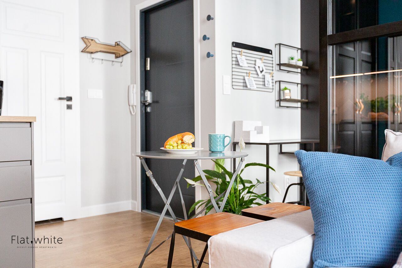 A modern living room with a sofa, a small table with a plate of food, a blue pillow, a plant, and a wall-mounted shelf with decorative items.