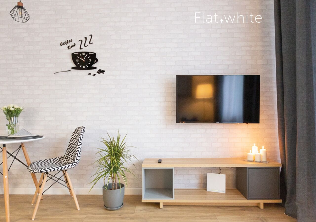 A modern living room with a flat-screen TV mounted on a brick wall, a black and white patterned chair, a small round table with a laptop, a potted plant, and a wooden console table with candles.