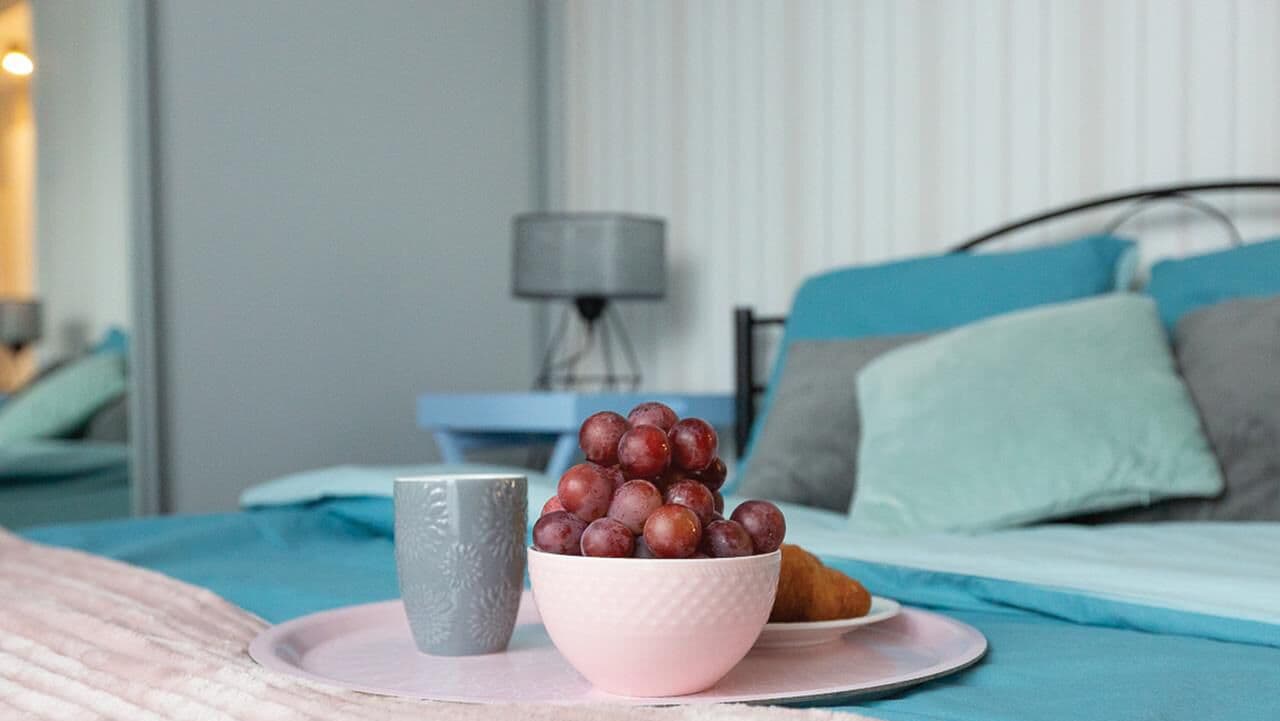 A neatly made bed with a blue bedspread, a bedside table with a lamp, and a bowl of grapes on a tray.