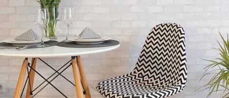 Un coin repas avec une table ronde dressée pour deux personnes, une chaise à motifs zigzag noir et blanc et une horloge murale au-dessus de la table.