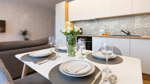 Un espace repas moderne avec une table blanche dressée pour deux personnes, un vase de fleurs blanches et une cuisine en arrière-plan.