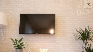 Un salon moderne avec un téléviseur à écran plat fixé à un mur en briques, une console en bois ornée de bougies, deux plantes en pot et deux tables rondes surmontées d’un tapis à motifs noir et blanc.