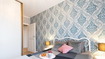 Un dormitorio con papel tapiz estampado, una cama blanca con múltiples almohadas, una mesita de noche con una lámpara y un plato de croissants sobre la cama.