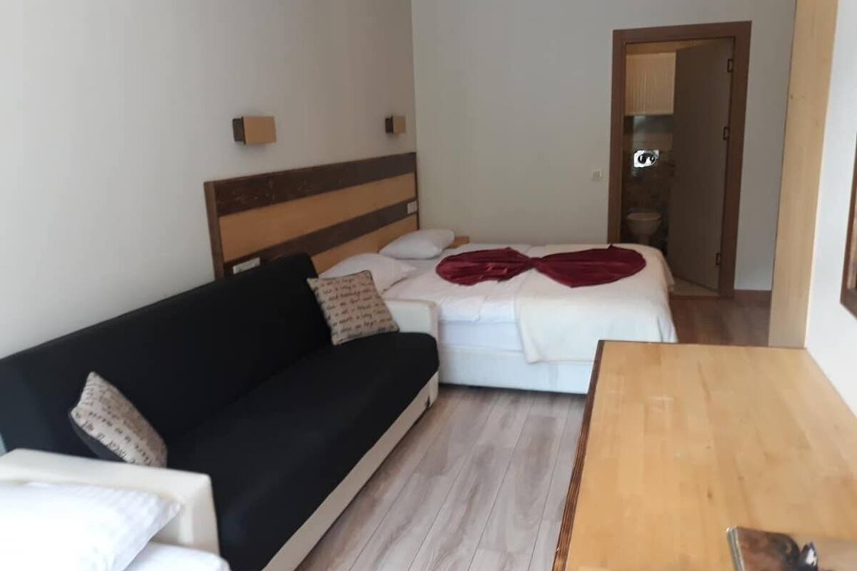 Quarto com sofá preto, cama branca com manta vermelha, mesa de madeira e um pequeno armário.