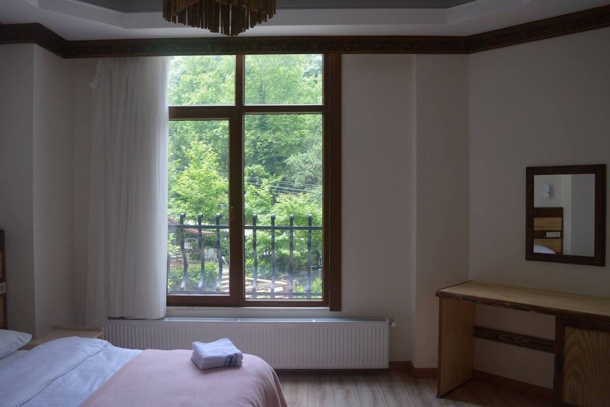 Quarto com uma grande janela, uma cama, uma mesa de cabeceira, um radiador e um espelho na parede.